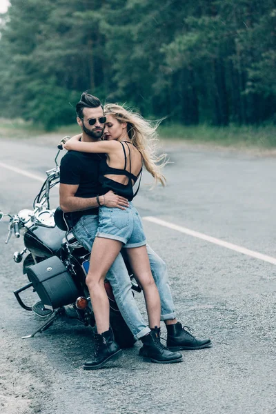 Young sexy couple of motorcyclists hugging near black motorcycle on road near green forest — Stock Photo