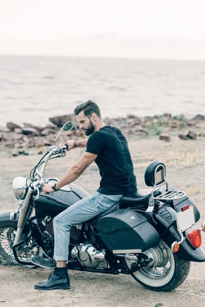 Bello motociclista su moto nero sulla spiaggia di sabbia vicino al mare — Foto stock