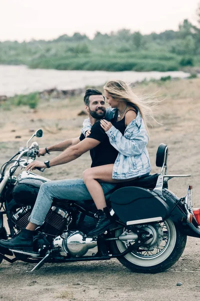 Sorrindo jovem casal de motociclistas abraçando na motocicleta preta na praia arenosa — Fotografia de Stock