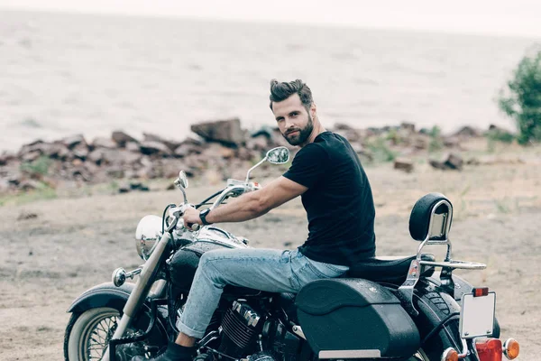 Guapo barbudo motociclista en moto negro en la playa de arena cerca del mar - foto de stock