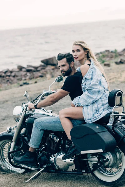 Joven pareja de ciclistas sentados en negro motocicleta en la playa cerca del río - foto de stock