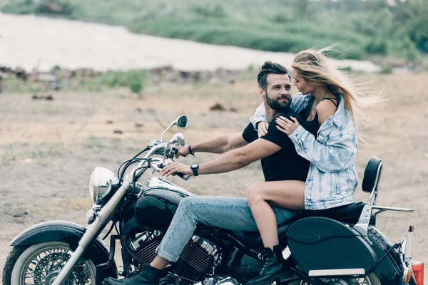 Jovem casal sorridente de motociclistas sentados em motocicleta preta na praia perto do rio — Fotografia de Stock