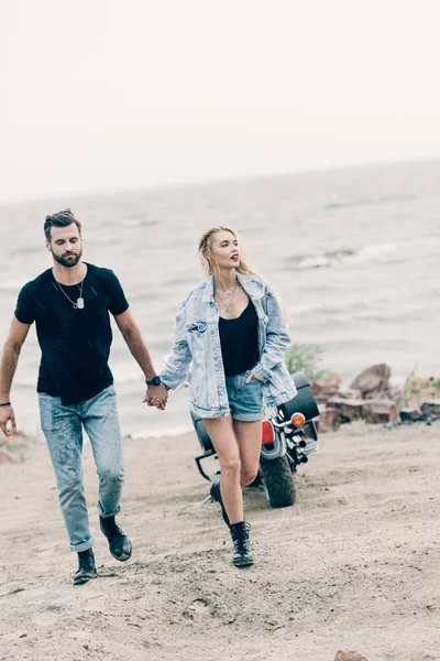 Jovem casal de motociclistas andando e de mãos dadas perto de motocicleta preta na praia arenosa — Fotografia de Stock