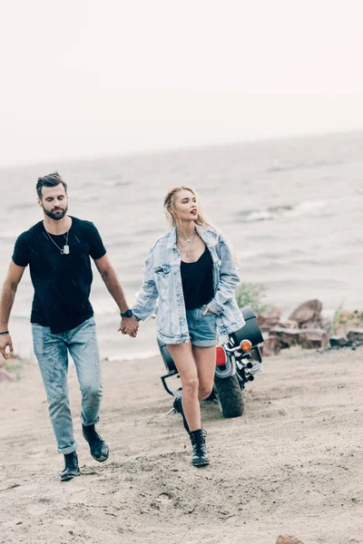 Joven pareja de ciclistas caminando y tomados de la mano cerca de la motocicleta negra en la playa de arena - foto de stock