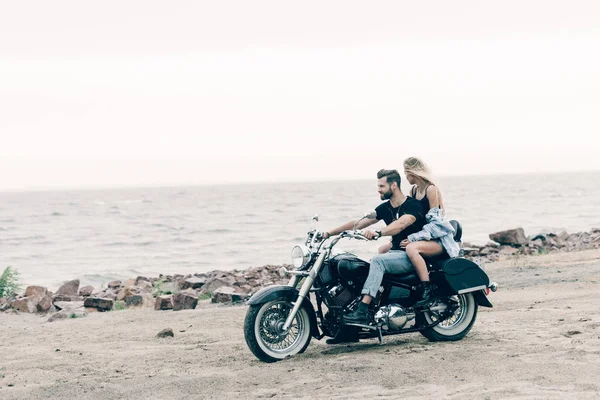 Молодая пара байкеров на черном мотоцикле на пляже возле реки — стоковое фото