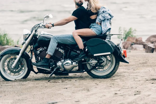 Обрізаний вид сексуальної молодої пари велосипедистів на чорному мотоциклі на піщаному пляжі — стокове фото