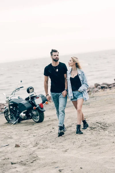 Joven pareja de ciclistas enamorados caminando y tomados de la mano cerca de la motocicleta negra en la playa de arena - foto de stock