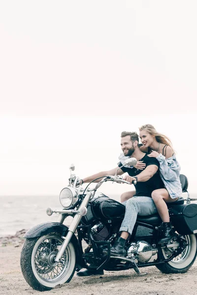 Happy young couple of bikers riding black motorcycle at sandy beach — Stock Photo