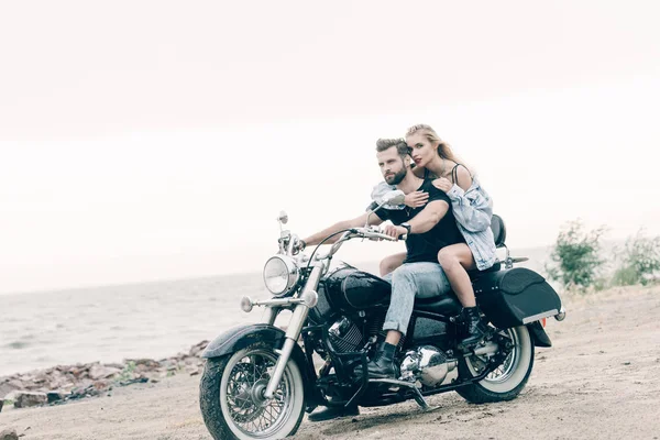 Молодая пара байкеров, катающихся на черном мотоцикле и обнимающихся на песчаном пляже — стоковое фото