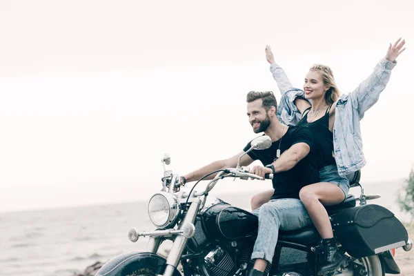Улыбающаяся молодая пара байкеров верхом на черном мотоцикле в то время как девушка поднимает руки в воздух возле реки — стоковое фото