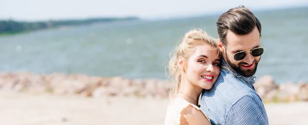 Felice donna bionda che abbraccia barbuto sorridente fidanzato in spiaggia, colpo panoramico — Foto stock