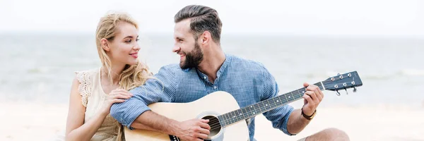 Улыбающаяся красивая молодая женщина сидит рядом с счастливым парнем с акустической гитарой на пляже у моря, панорамный снимок — стоковое фото