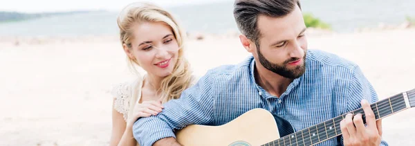 Sonriente hermosa joven sentada cerca de novio con guitarra acústica en la playa cerca del mar, tiro panorámico - foto de stock