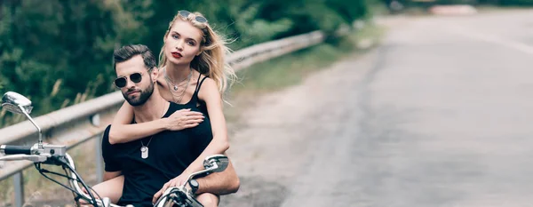 Jeune couple de motards brutaux étreignant sur la moto noire sur la route près de la forêt verte, vue panoramique — Photo de stock