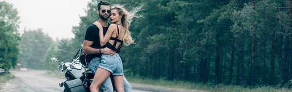 Jovem casal sexy de motociclistas abraçando perto de motocicleta preta na estrada perto de floresta verde, tiro panorâmico — Fotografia de Stock