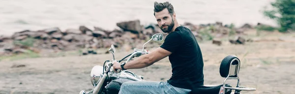 Handsome bearded motorcyclist on black motorcycle at sandy beach near sea, panoramic shot — Stock Photo