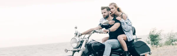 Joven amante pareja de ciclistas sentado en negro motocicleta en la playa cerca del río, tiro panorámico - foto de stock