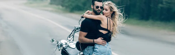 Jovem casal sexy de motociclistas abraçando perto de motocicleta preta na estrada perto de floresta verde, tiro panorâmico — Fotografia de Stock