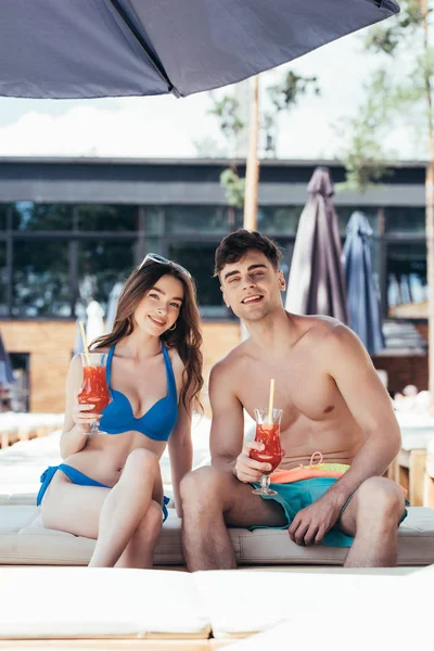 Alegre joven pareja sentado en cubierta silla con vasos de bebida refrescante y mirando a la cámara - foto de stock