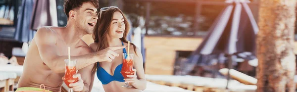 Plano panorámico de joven alegre señalando con el dedo mientras está sentado cerca de novia bonita con vasos de bebida refrescante - foto de stock
