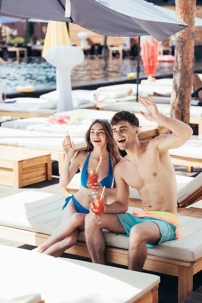 Весела молода пара махає руками, сидячи на стільці на палубі з келихами освіжаючого напою — стокове фото