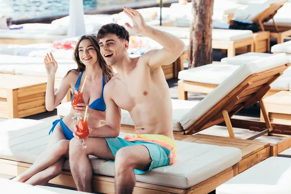 Feliz joven pareja sosteniendo vasos de bebida refrescante y agitando las manos mientras mira hacia otro lado - foto de stock