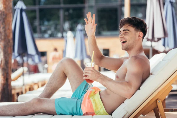 Cheerful young man waving looking away and waving hand while relaxing on chaise lounge with glass of refreshing drink — Stock Photo