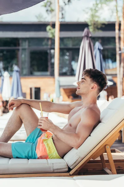 Мечтательный молодой человек смотрит в сторону, расслабляясь в шезлонге с бокалом освежающего напитка — стоковое фото