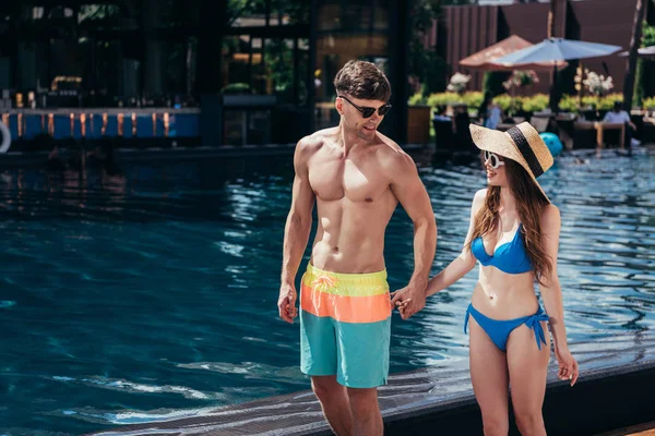 Heureux jeune couple dans des lunettes de soleil tenant la main tout en se tenant près de la piscine — Photo de stock