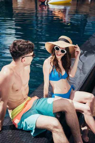 Alegre joven pareja hablando y sonriendo mientras se sienta cerca de la piscina - foto de stock