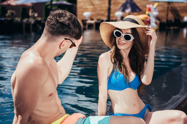 Attraktive junge Frau mit Sonnenbrille und Strohhut am Pool in der Nähe ihres Freundes — Stockfoto