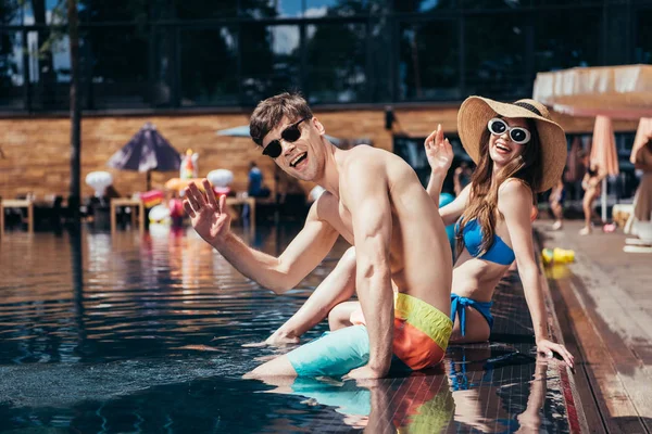 Fröhliches junges Paar mit Sonnenbrille winkt in die Kamera, während es sich am Pool entspannt — Stockfoto