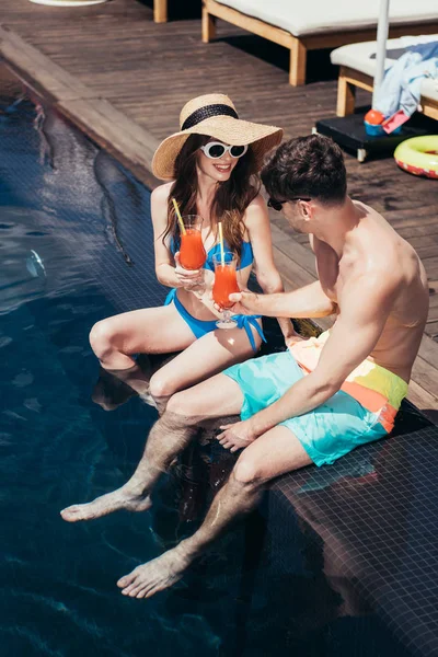 Щаслива молода пара смердить окуляри з освіжаючим напоєм, сидячи на узбережжі басейну — стокове фото