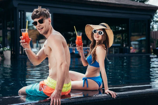 Fröhliches junges Paar lächelt in die Kamera, während es am Pool mit Gläsern erfrischenden Getränks sitzt — Stockfoto