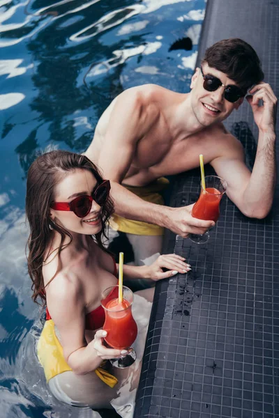 Feliz joven pareja en gafas de sol sosteniendo vasos de bebida refrescante y sonriendo a la cámara - foto de stock