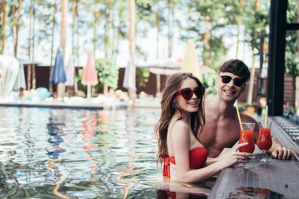 Feliz pareja joven relajarse en la piscina con glases de bebida refrescante una sonrisa en la cámara - foto de stock