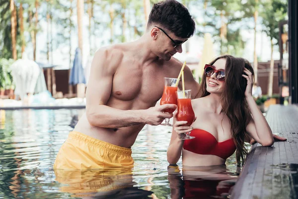 Веселая молодая пара, звенящие стаканы освежающего напитка во время отдыха в бассейне — стоковое фото