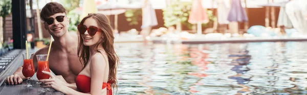 Panoramic shot of happy young couple relaxing in swimming pool with glasses of refreshing drink — Stock Photo