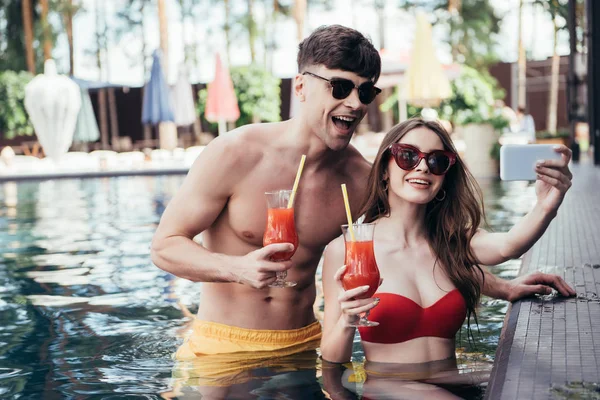 Alegre pareja joven tomando selfie mientras se relaja en la piscina con vasos de bebida refrescante - foto de stock