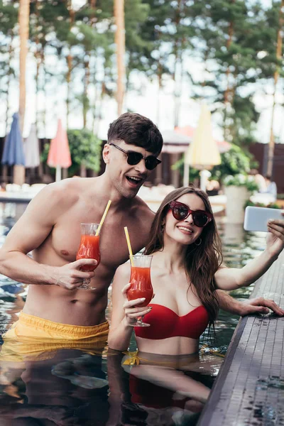 Весела пара тримає келихи освіжаючого напою і приймає селфі під час відпочинку в басейні — стокове фото