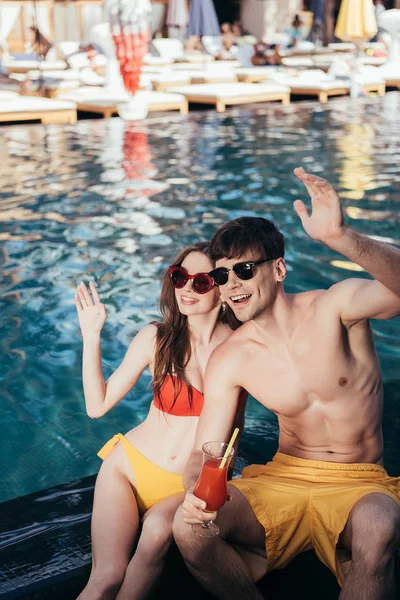 Весела молода пара дивиться в сторону і махає руками, сидячи біля басейну — стокове фото