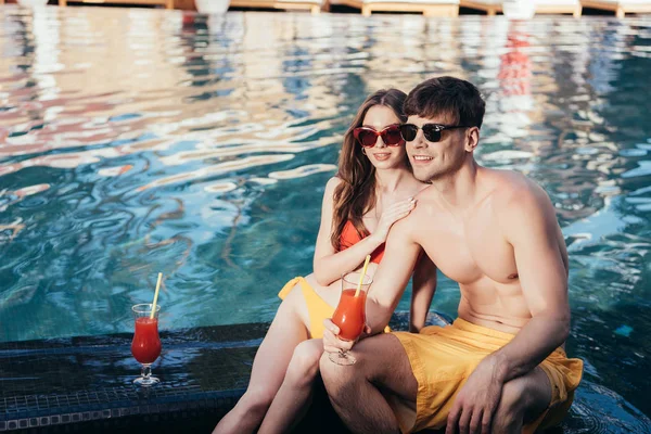 Счастливая молодая пара смотрит в сторону, сидя у бассейна с бокалами освежающего напитка — стоковое фото