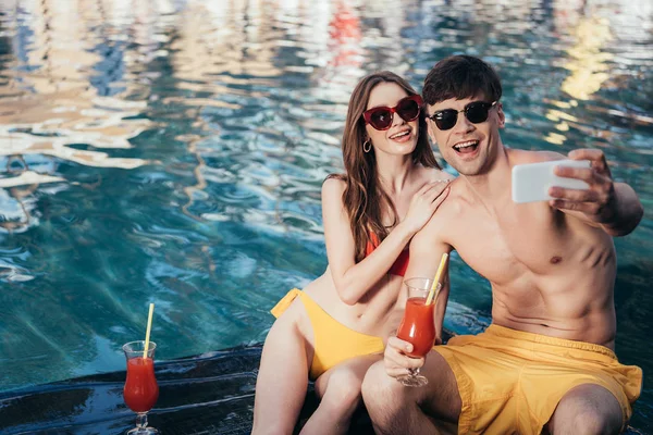 Joyeux jeune couple prenant selfie tout en étant assis au bord de la piscine avec des verres de boisson rafraîchissante — Photo de stock