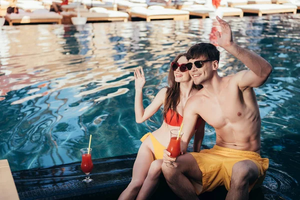 Счастливая молодая пара смотрит в сторону и машет руками, сидя у бассейна — стоковое фото