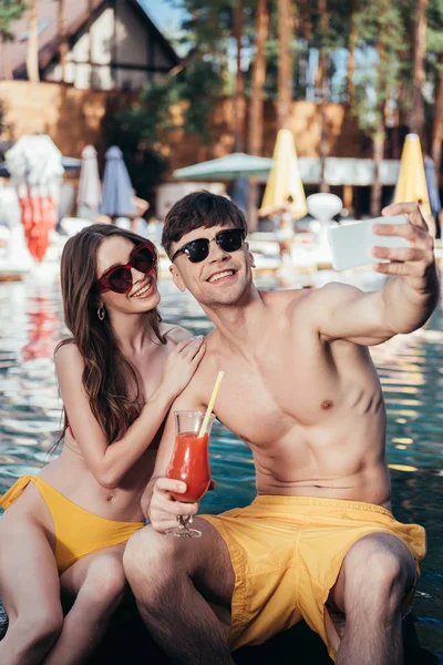 Joyeux jeune couple prenant selfie tout en étant assis au bord de la piscine avec des verres de boisson rafraîchissante — Photo de stock