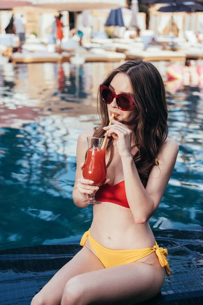 Bastante joven mujer en traje de baño y gafas de sol beber bebida refrescante mientras está sentado en la piscina - foto de stock