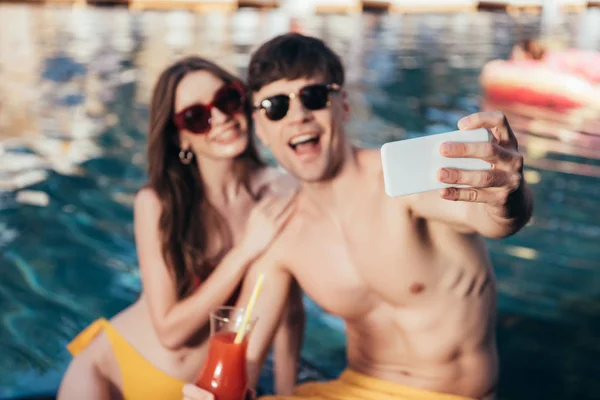 Foco seletivo de jovem casal alegre tomando selfie enquanto sentado à beira da piscina — Fotografia de Stock