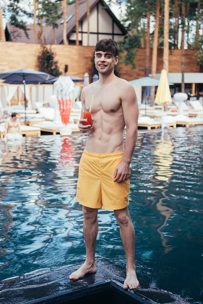Красивий молодий чоловік посміхається на камеру, стоячи біля басейну зі склянкою освіжаючого напою — стокове фото