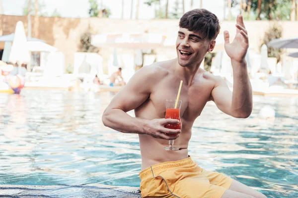 Веселий молодий чоловік, дивлячись і махаючи рукою, відпочиває біля басейну зі склянкою освіжаючого напою — стокове фото