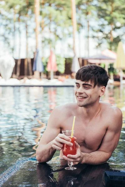 Joven guapo mirando hacia otro lado mientras se relaja en la piscina con un vaso de bebida refrescante - foto de stock